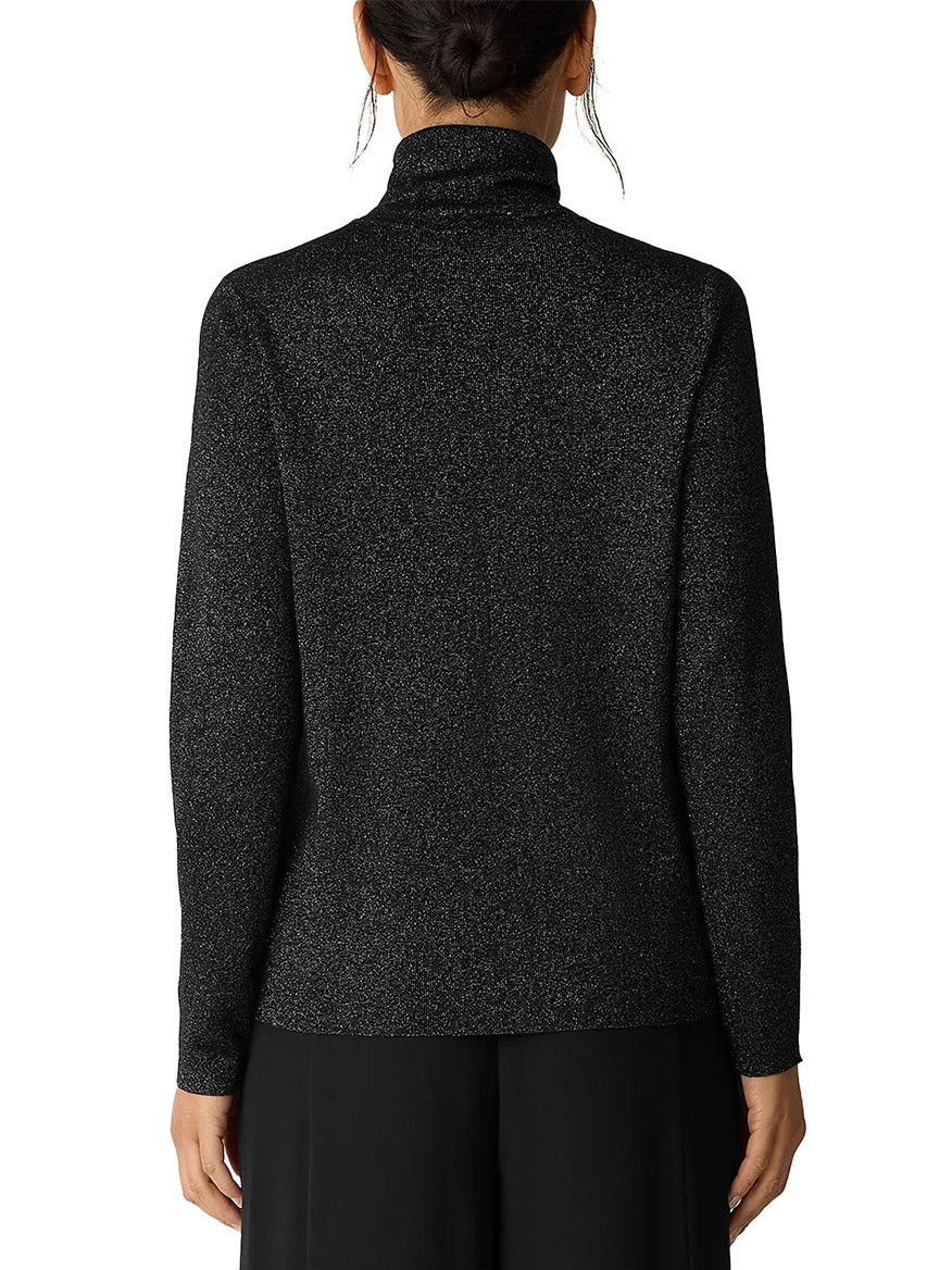 A person is seen from behind wearing the Eileen Fisher Wool Lurex Turtleneck Sweater in Black, featuring subtle metallic shimmer, paired with black pants.