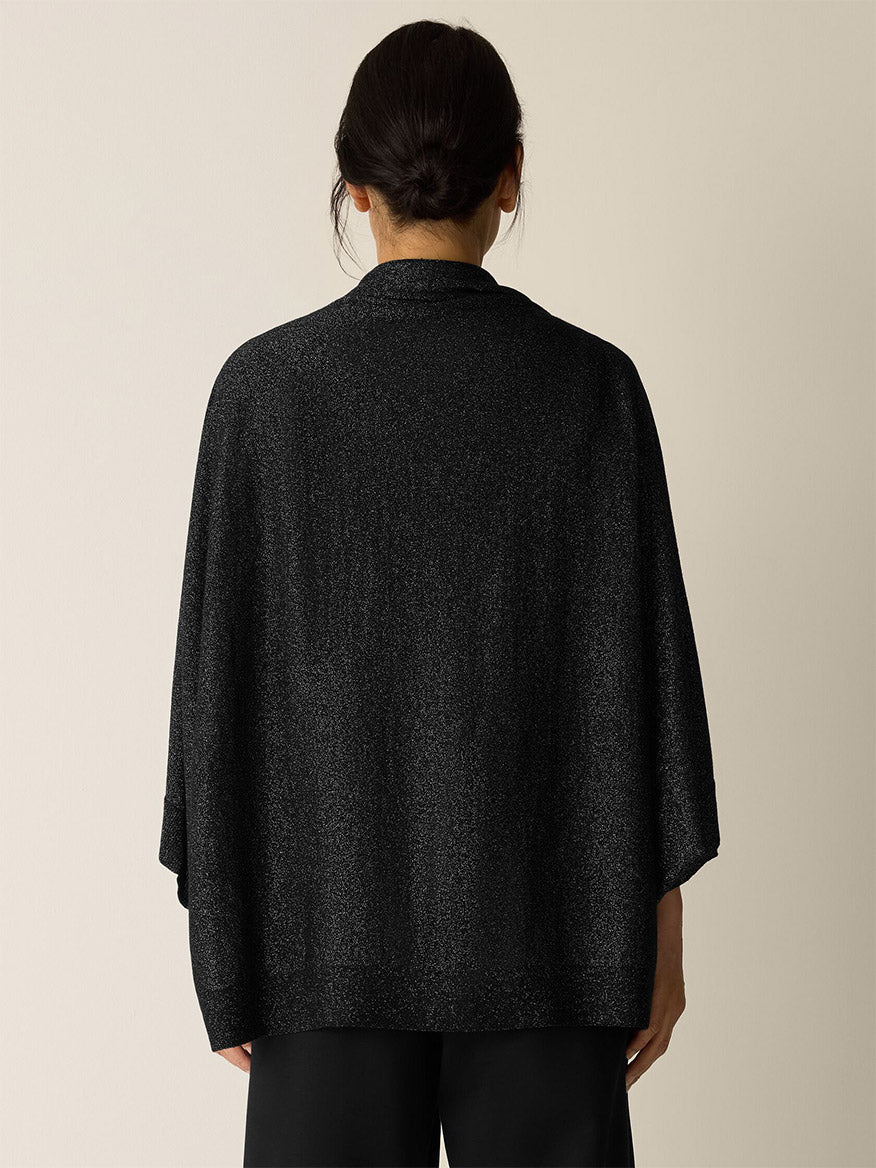 A person stands facing away, wearing the Eileen Fisher Wool Shimmer Serape in Black with wide sleeves and black pants against a neutral background.