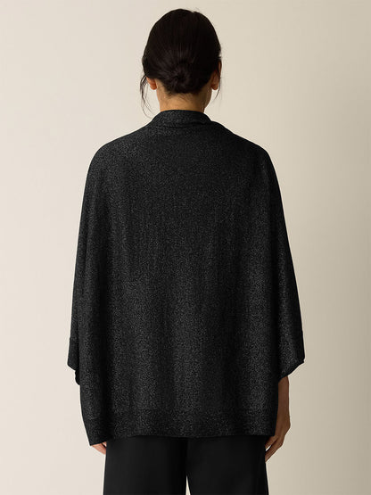 A person stands facing away, wearing the Eileen Fisher Wool Shimmer Serape in Black with wide sleeves and black pants against a neutral background.