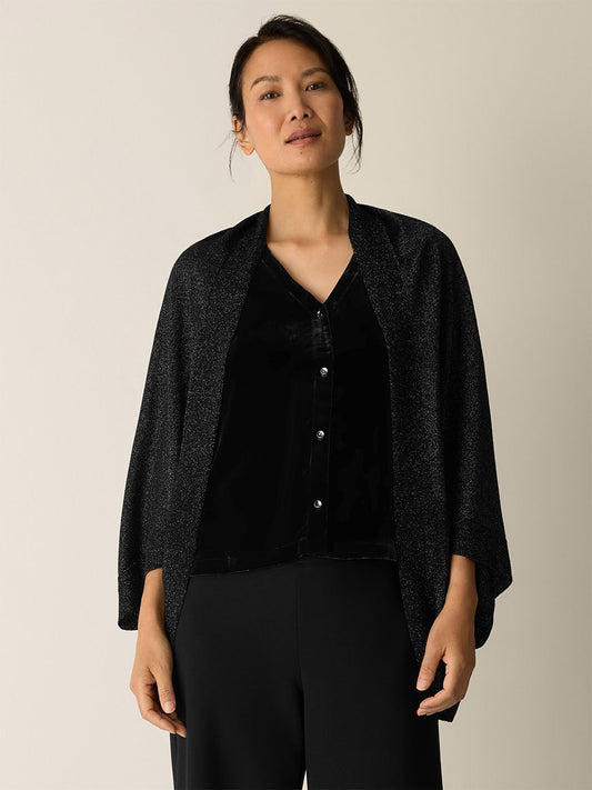 A person wearing the Eileen Fisher Wool Shimmer Serape in Black over a black button-up top and black pants stands against a plain light background.