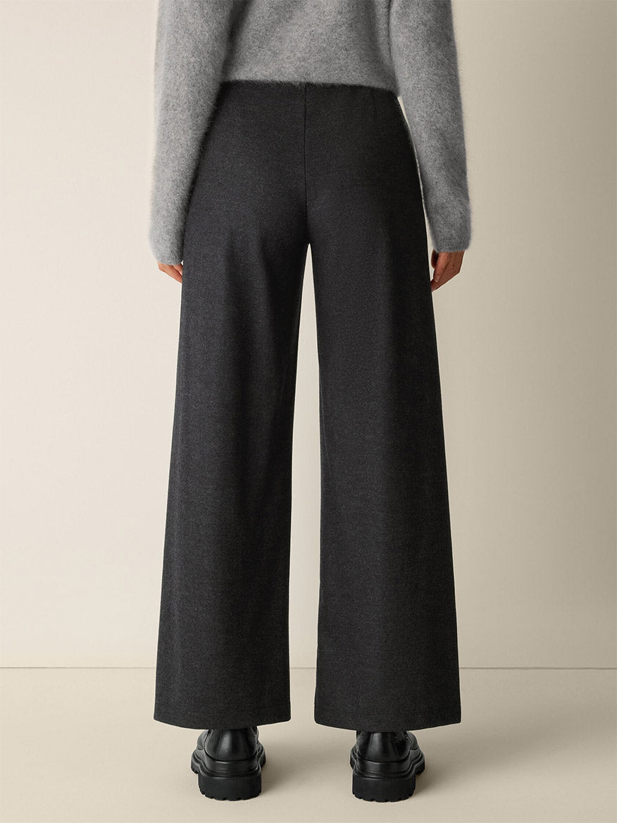Model wearing Eileen Fisher Mélange Boiled Wool Jersey Wide-Leg Pant in charcoal, chunky black shoes, and a light gray fuzzy sweater, shown from the back against a plain beige background.