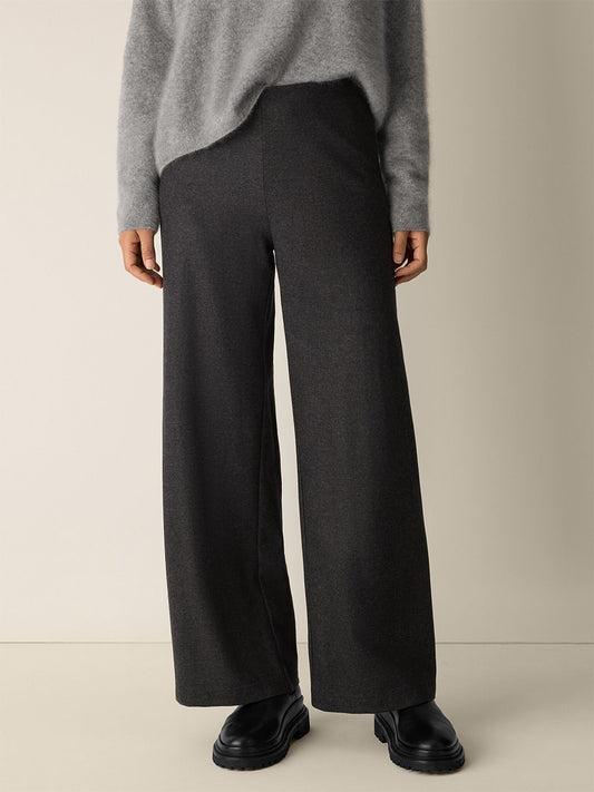 A person stands against a plain light background wearing a grey long-sleeve sweater, black shoes, and Eileen Fisher Mélange Boiled Wool Jersey Wide-Leg Pant in Charcoal.