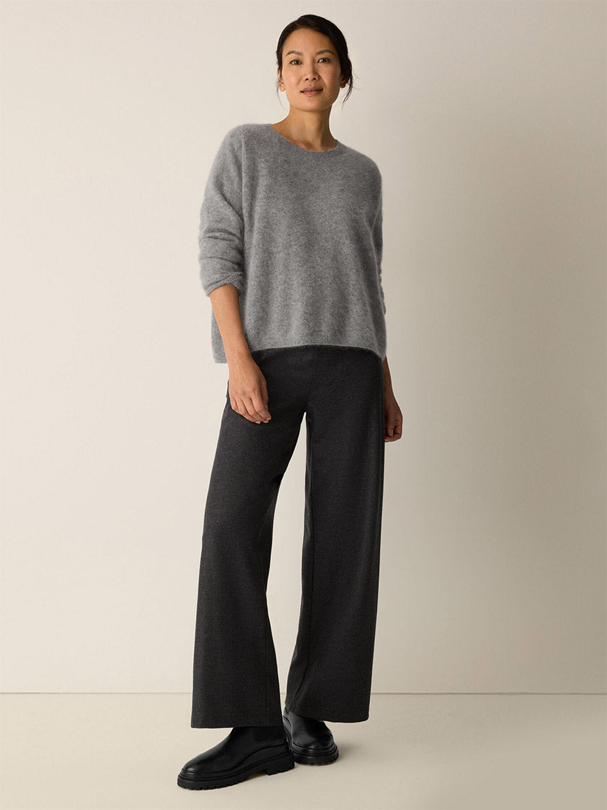 A woman stands against a plain light wall, wearing a loose grey sweater, Eileen Fisher Mélange Boiled Wool Jersey Wide-Leg Pant in Charcoal, and black shoes.