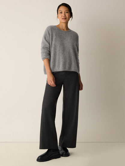 A woman stands against a plain light wall, wearing a loose grey sweater, Eileen Fisher Mélange Boiled Wool Jersey Wide-Leg Pant in Charcoal, and black shoes.