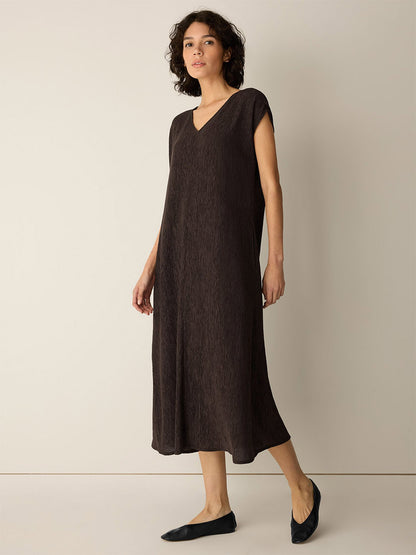 A woman stands against a plain light wall, wearing the Eileen Fisher Woven Plissé V-Neck Dress in Truffle—a loose-fitting, dark brown midi dress with short sleeves—paired with black flats.