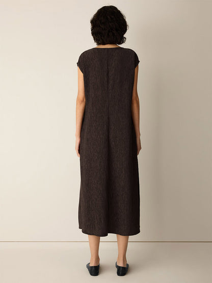 A person stands indoors facing away, wearing the Eileen Fisher Woven Plissé V-Neck Dress in Truffle—a short-sleeve, calf-length dark brown dress—paired with black flats against a plain light-colored wall and floor.