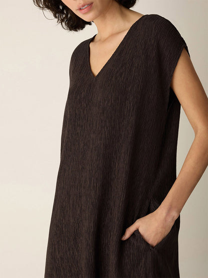 A woman stands against a light background, one hand in her pocket, wearing the Eileen Fisher Woven Plissé V-Neck Dress in Truffle. The dress features a loose fit, textured weave, short sleeves, and a V-neckline. Her face is partially out of the frame.