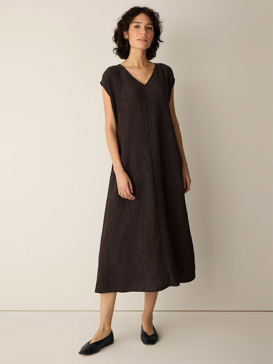 A woman wearing the Eileen Fisher Woven Plissé V-Neck Dress in Truffle and black flats, standing against a plain beige background.