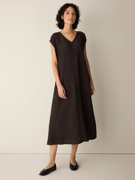 A woman wearing the Eileen Fisher Woven Plissé V-Neck Dress in Truffle and black flats, standing against a plain beige background.
