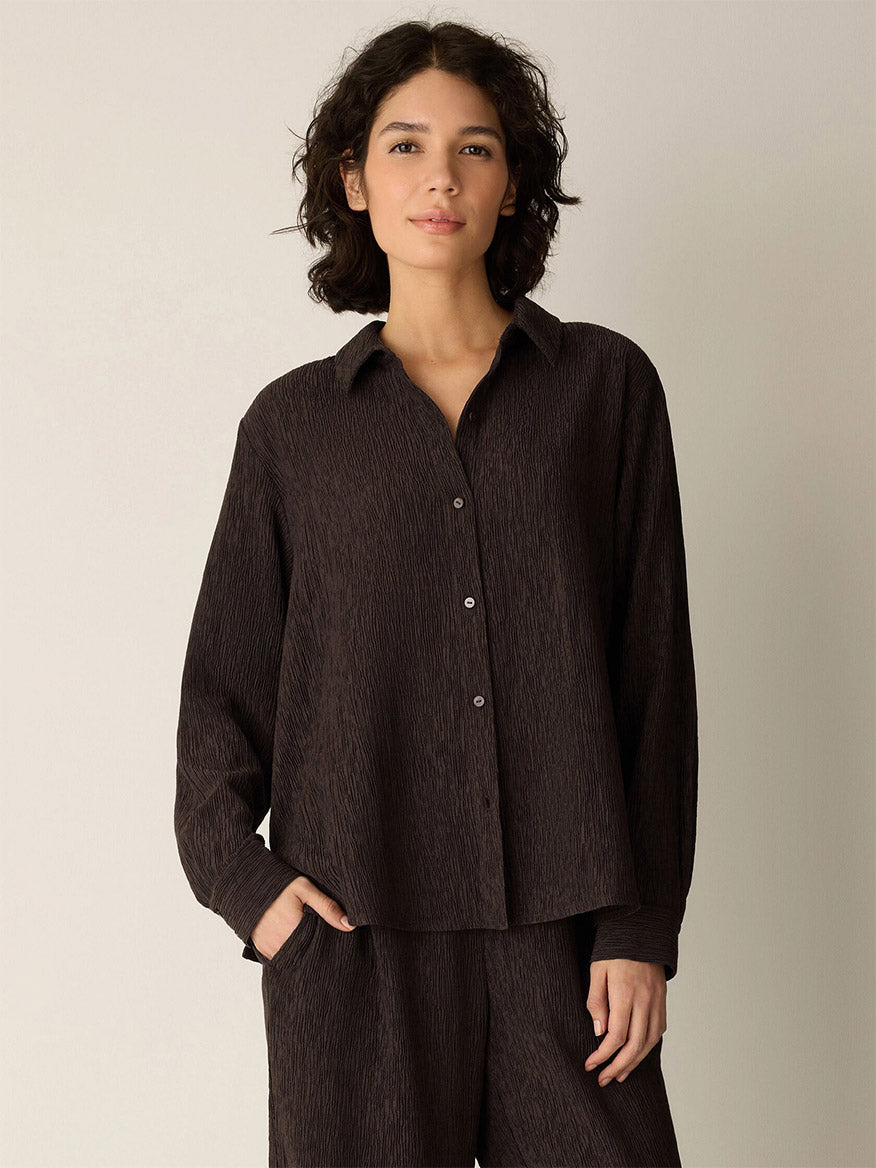 A person with curly dark hair wears the Eileen Fisher Woven Plissé Classic Collar Shirt in Truffle, styled loosely with matching textured pants, standing against a plain light background.