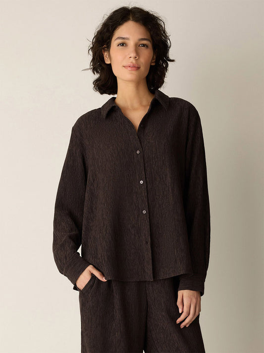 A person with curly dark hair wears the Eileen Fisher Woven Plissé Classic Collar Shirt in Truffle, styled loosely with matching textured pants, standing against a plain light background.
