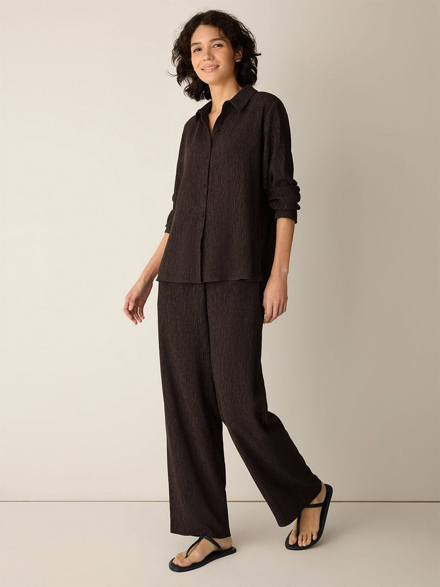A person stands against a neutral wall, wearing the loose-fitting Eileen Fisher Woven Plissé Classic Collar Shirt in Truffle with matching pants and flip-flops.
