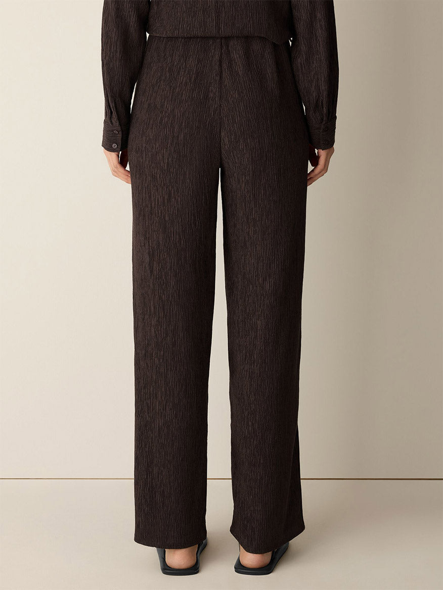 A person stands against a neutral background, shown from the back, wearing the Eileen Fisher Woven Plissé Straight Pant in Truffle paired with a matching drapey top and black pointed-toe shoes.