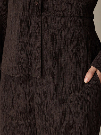 Close-up of a person wearing the Eileen Fisher Woven Plissé Straight Pant in Truffle, paired with a brown textured shirt featuring a drapey weave, with one hand partially tucked into the pocket.