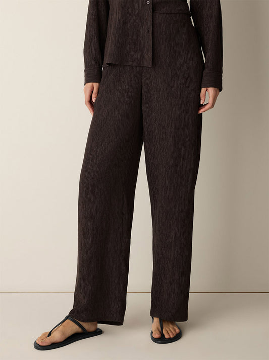 Wearing the Eileen Fisher Woven Plissé Straight Pant in Truffle—loose, dark brown textured straight pants with a drapey weave—paired with a matching long-sleeve top and black sandals, standing against a plain beige wall.