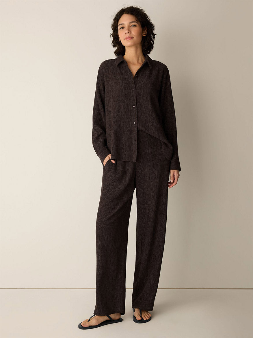 A person stands against a plain wall wearing a loose dark brown button-up shirt crafted from a drapey weave, the Eileen Fisher Woven Plissé Straight Pant in Truffle, and black sandals.
