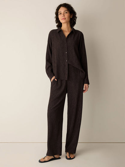A person stands against a plain wall wearing a loose dark brown button-up shirt crafted from a drapey weave, the Eileen Fisher Woven Plissé Straight Pant in Truffle, and black sandals.