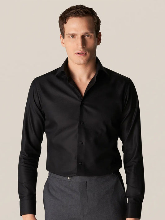 A person is seen wearing an Eton Slim Fit Black Textured Twill Dress Shirt paired with gray pants, exemplifying classic business attire against a simple backdrop.