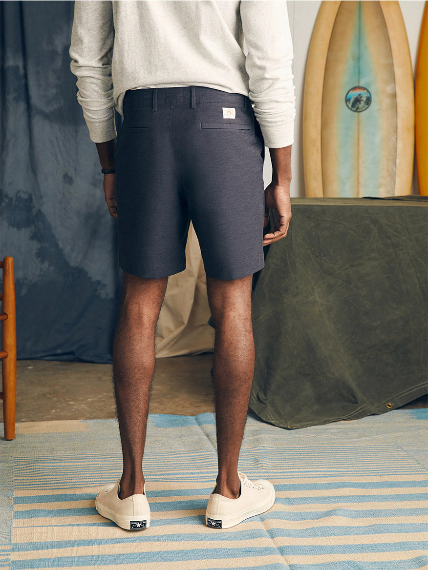 A man standing in a casual setting wearing navy Faherty Brand All Day Shorts in Charcoal and slip-on shoes with a longboard surfboard in the background.