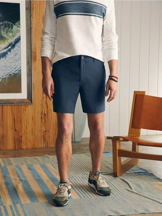 Man wearing a striped sweatshirt and navy Faherty Brand All Day Shorts in Dark Blue Nights designed with eco-friendly fabric, standing beside a wooden chair in a room with coastal decor.
