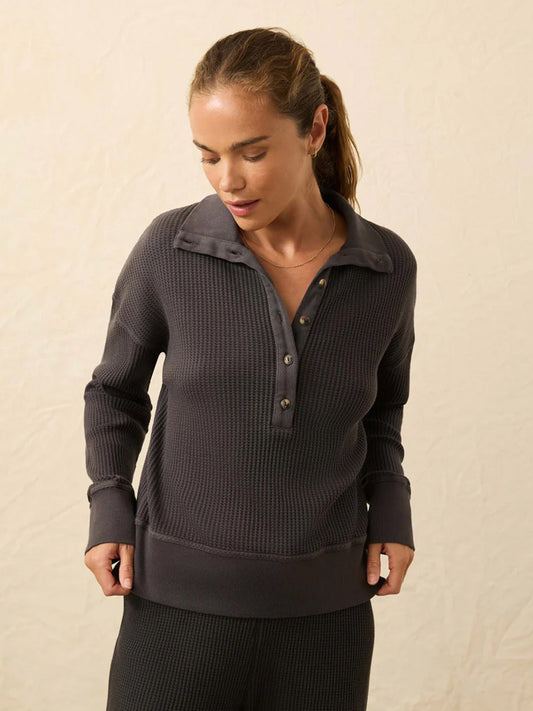 A woman wears the Faherty Brand Après Waffle Henley in Washed Black—a relaxed-fit, waffle-textured top with buttons—as she stands against a plain light background, looking down.