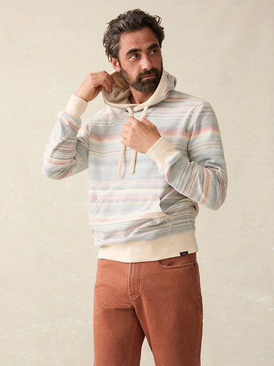 A bearded man wearing a striped, stylish sweatshirt and rust-colored pants adjusts the hood of his Faherty Brand Byron Bay Hoodie in Island Sunrise.