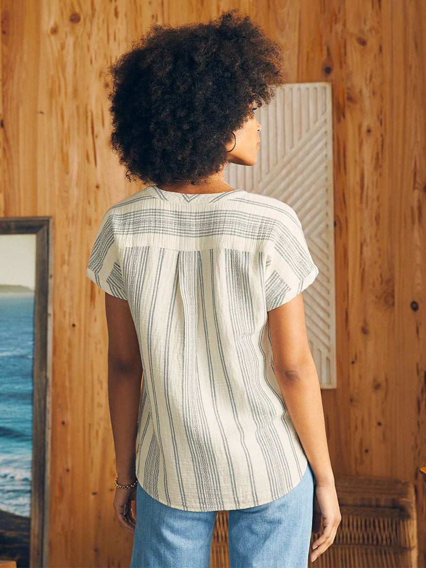 Woman standing indoors facing away from the camera, wearing a Faherty Brand Dream Cotton Gauze Desmond Top in Cream Tidal Wave Dobby and denim jeans, with a backdrop of wood paneling and a view of the sea through a window