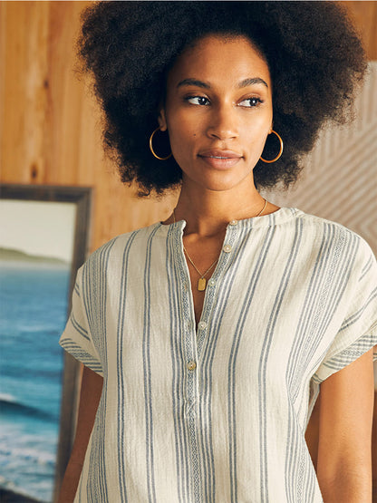 A woman with an afro wearing a Faherty Brand Dream Cotton Gauze Desmond Top in Cream Tidal Wave Dobby and hoop earrings, looking away thoughtfully.