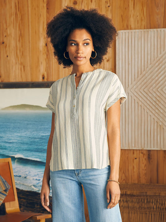 Woman in a Faherty Brand Dream Cotton Gauze Desmond Top in Cream Tidal Wave Dobby and jeans standing in front of a wood panel background with a beach painting.