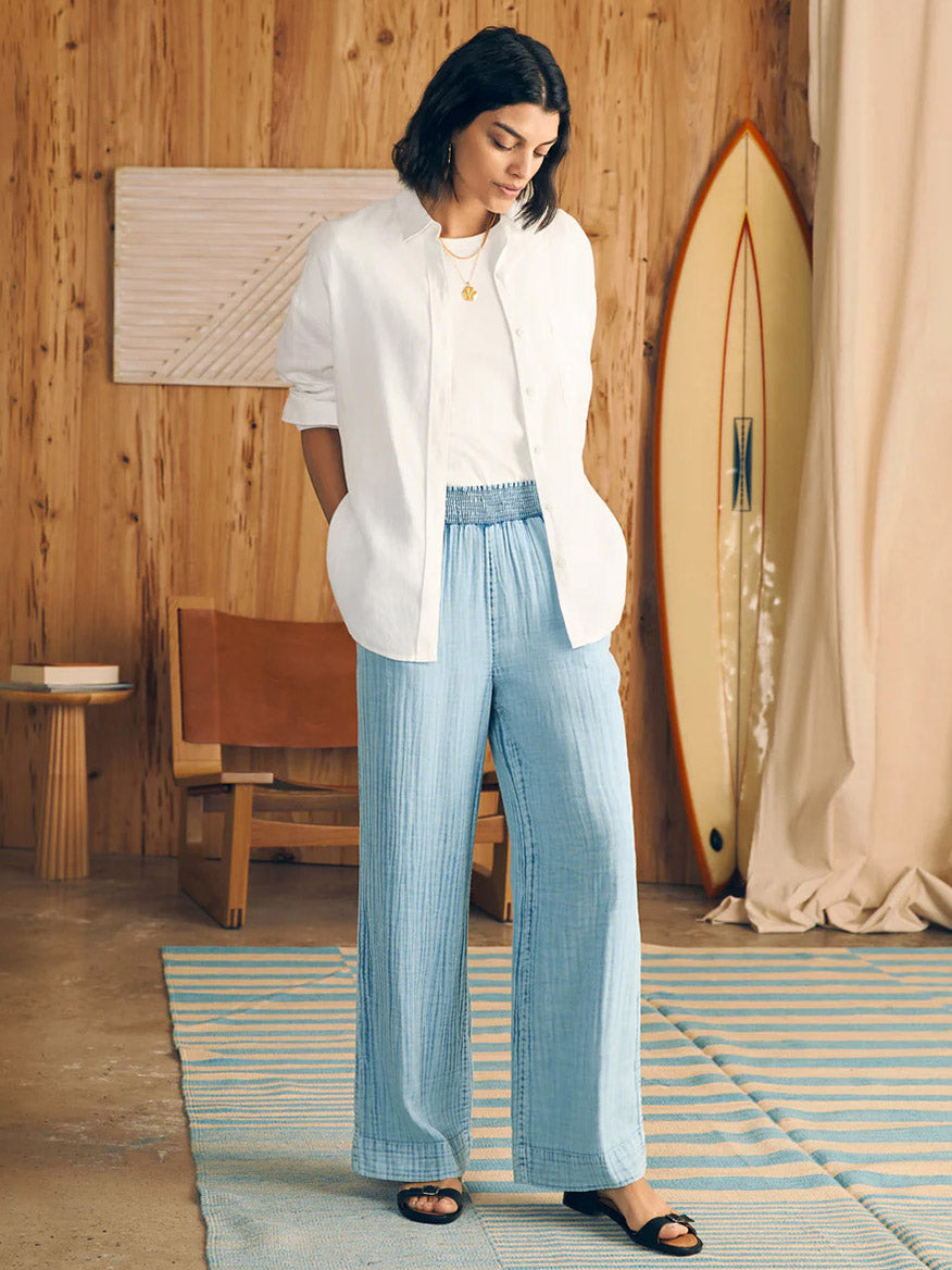 A woman standing in a wooden room, dressed in a Faherty Brand Dream Cotton Gauze Wide Leg Pant in Light Indigo Wash and a casual white shirt, next to a surfboard leaning against the wall.