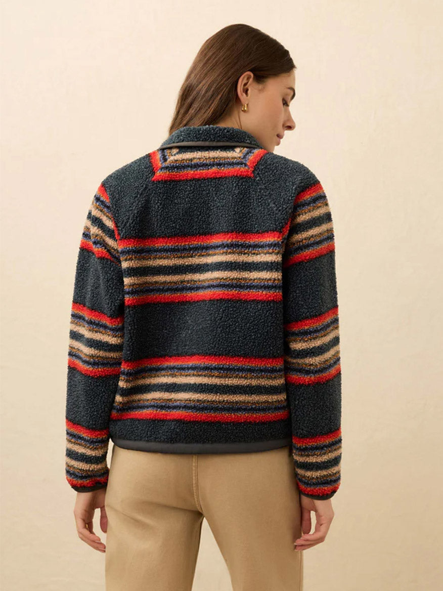 Someone with long brown hair wears the Faherty Brand Fireside Fleece Popover in Fireside Stripe, featuring red, tan, and beige horizontal stripes, paired with beige pants against a plain light background.