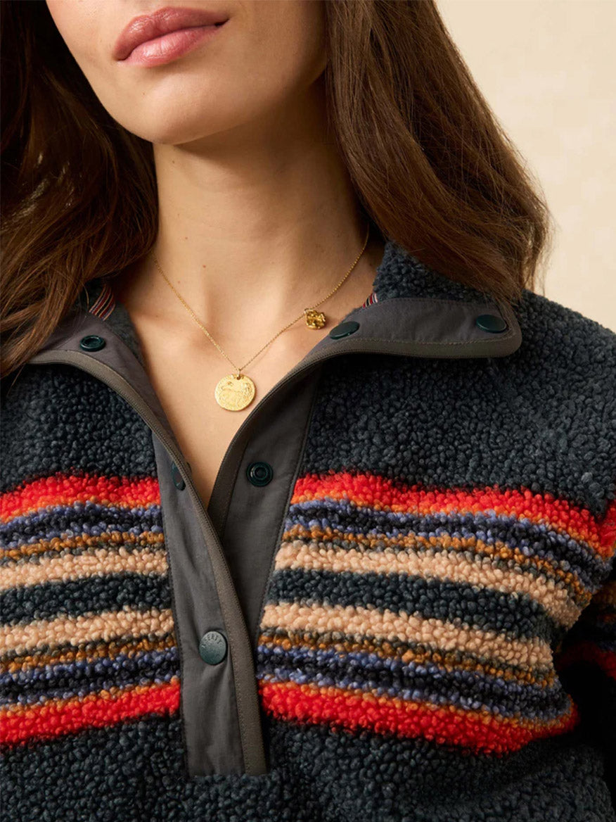 A woman models the Faherty Brand Fireside Fleece Popover in Fireside Stripe, a cozy textured pullover in red, tan, and navy, paired with two gold pendant necklaces for sporty warmth and style.