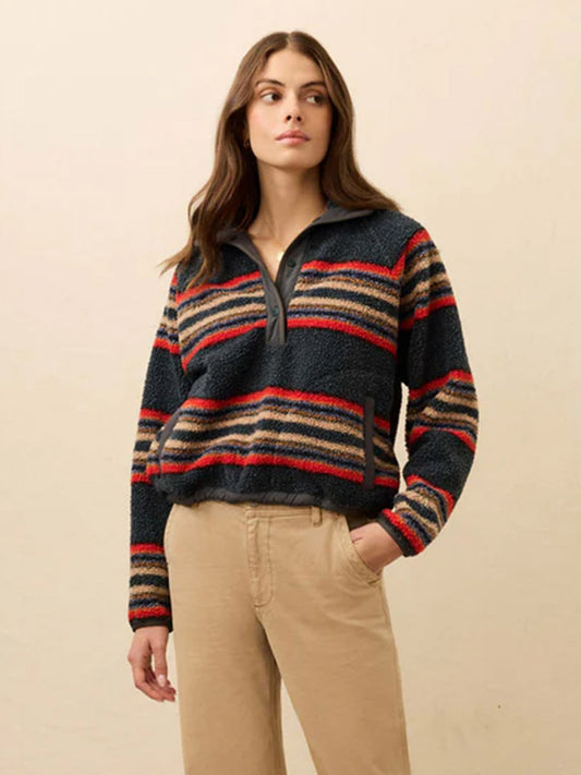 A woman wearing the Faherty Brand Fireside Fleece Popover in Fireside Stripe—cozy with a quarter-zip collar—paired with beige pants, stands against a plain light background.
