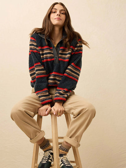 A person sits on a wooden stool against a light background, wearing the Faherty Brand Fireside Fleece Popover in Fireside Stripe for cozy warmth, along with beige pants, patterned socks, and black sneakers.
