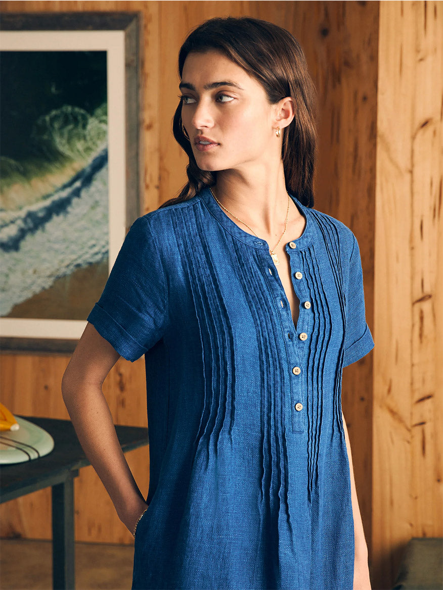Woman in a Faherty Brand Gemina Basketweave Dress in Indigo standing beside a wooden wall with a framed picture in the background.