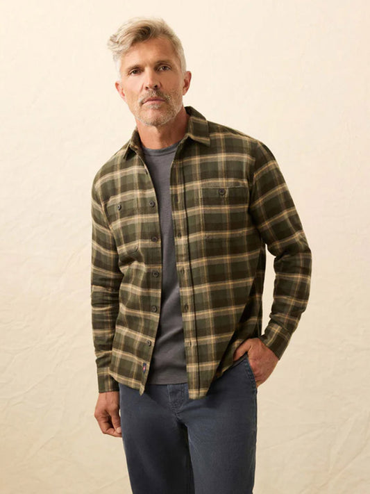 A man with gray hair wears the Faherty Brand Heirloom Portuguese Flannel Shirt in Garrett Verde Plaid over a gray t-shirt and blue jeans, standing against a plain light background.