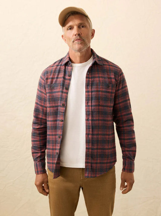 A man wearing a beige cap, tan pants, and a Faherty Brand Heirloom Portuguese Flannel Shirt in Porto Covo Plaid over a white t-shirt stands against a neutral background.