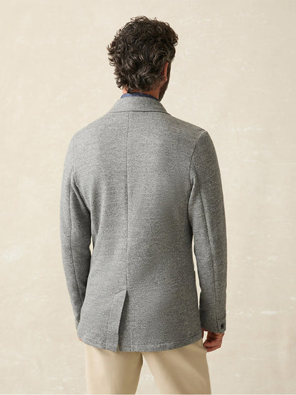 A man with curly hair, standing with his back to the camera, is wearing the Faherty Brand Inlet Knit Blazer in Medium Grey Melange and light-colored pants against a neutral background.