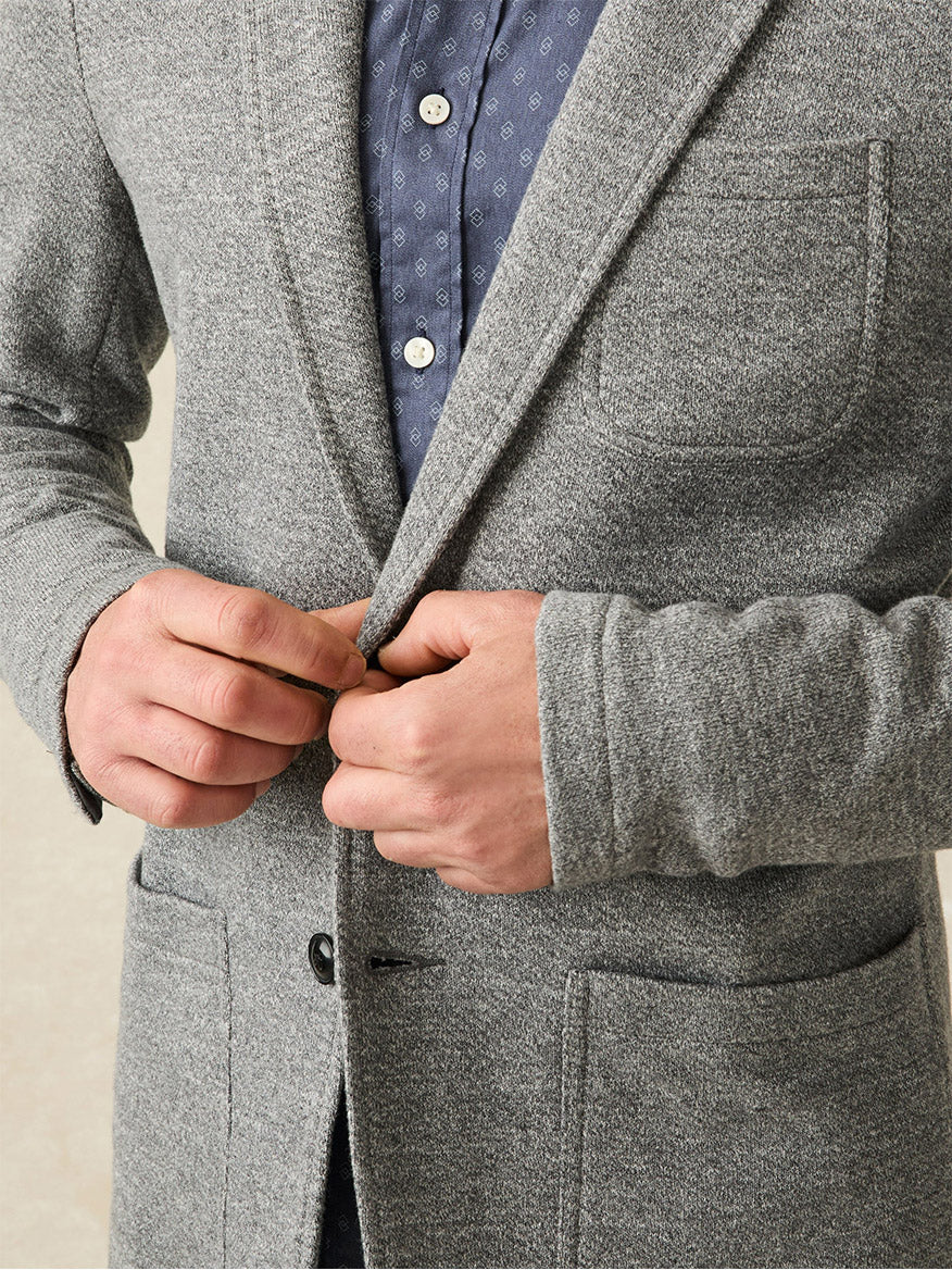 Close-up of a person buttoning the Faherty Brand Inlet Knit Blazer in Medium Grey Melange over a blue patterned shirt, with a focus on their hands and the front of the blazer.