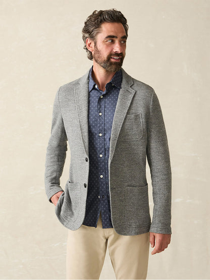 A man with dark hair and a beard wears a Faherty Brand Inlet Knit Blazer in Medium Grey Melange over a patterned blue shirt and beige pants, looking to the side.