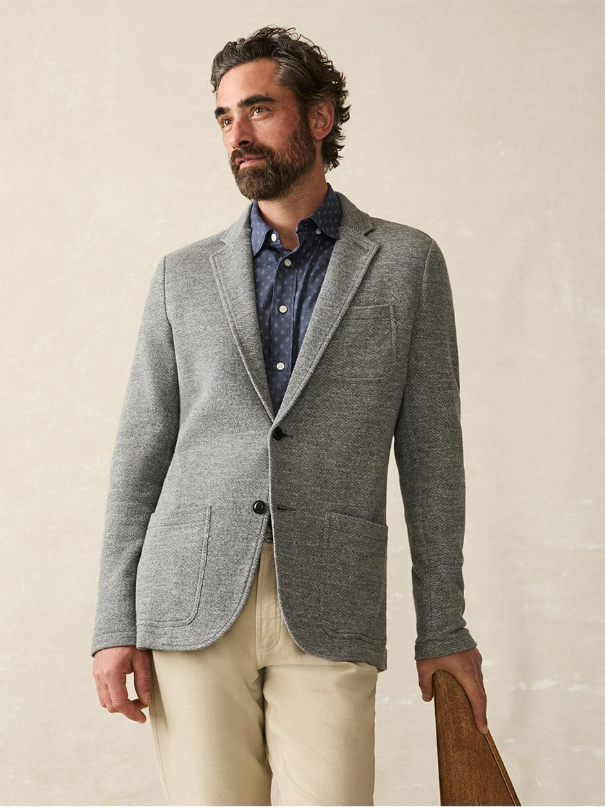 A bearded man in a Faherty Brand Inlet Knit Blazer in Medium Grey Melange over a blue shirt and beige pants stands against a neutral background, holding a wooden object.