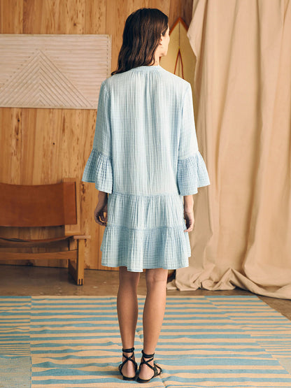 Woman standing in a room facing away from the camera, wearing a Faherty Brand Dream Cotton Gauze Kasey Dress in Light Indigo Wash, featuring long billowy sleeves and a tiered mini skirt, paired with black str