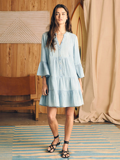Woman in a Faherty Brand Dream Cotton Gauze Kasey Dress in Light Indigo Wash with long billowy sleeves, standing in a room with wood paneling and a surfboard in the background.