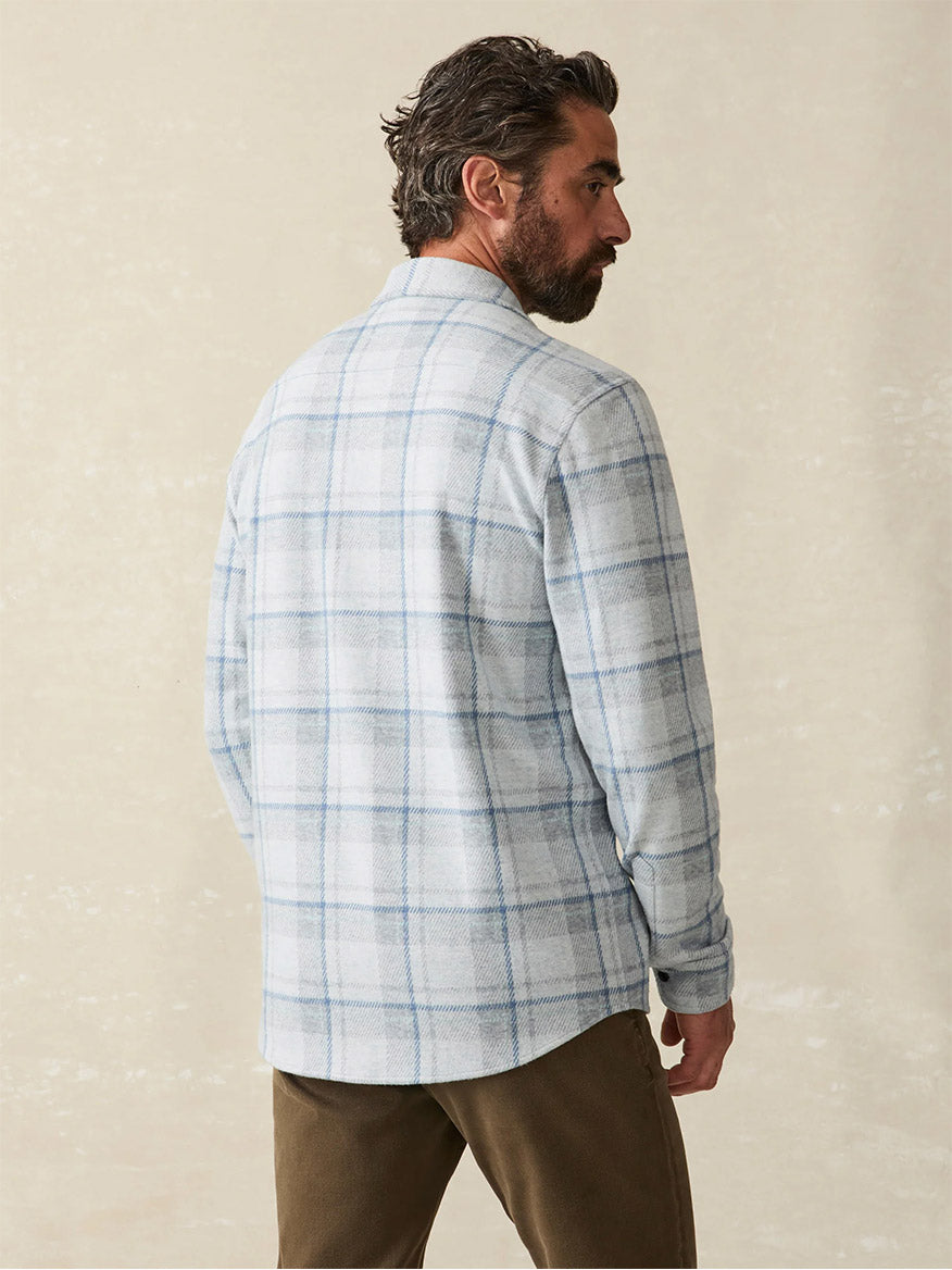 A man with a beard, wearing the Faherty Brand Legend Sweater Shirt in Maine Sky Plaid, stands facing away to showcase the impeccable design of this bestselling shirt. The soft knit fabric and subtle four-way stretch ensure ultimate comfort and style.