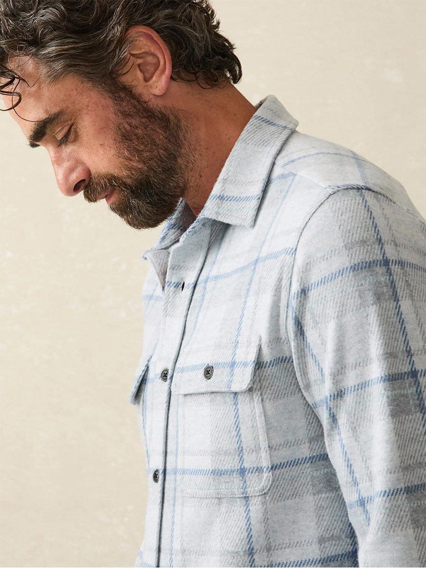 A bearded man with wavy hair is looking downwards, wearing the Faherty Brand Legend Sweater Shirt in Maine Sky Plaid against a plain background. This bestselling shirt features a soft knit fabric for ultimate comfort.