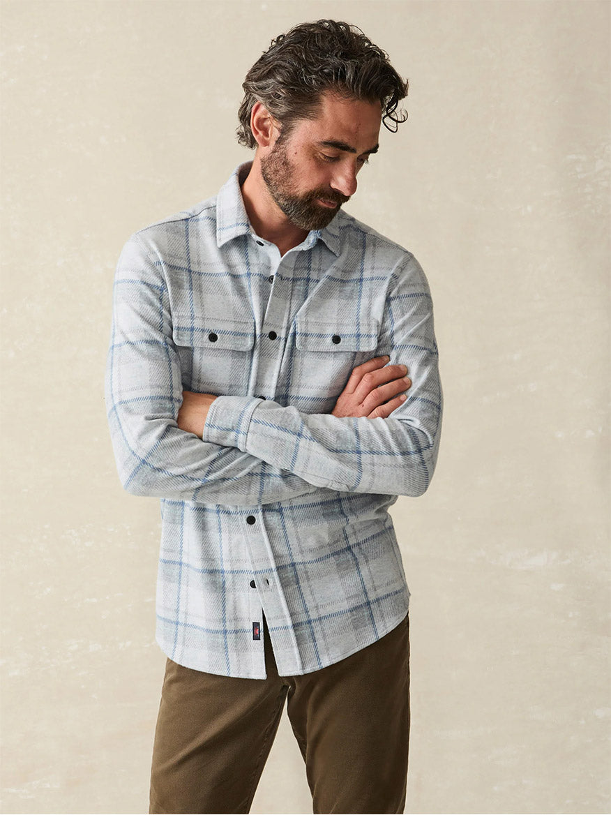 A man with dark hair and a beard, dressed in Faherty Brand Legend Sweater Shirt in Maine Sky Plaid and brown pants made from soft knit fabric, stands with arms crossed, looking down.