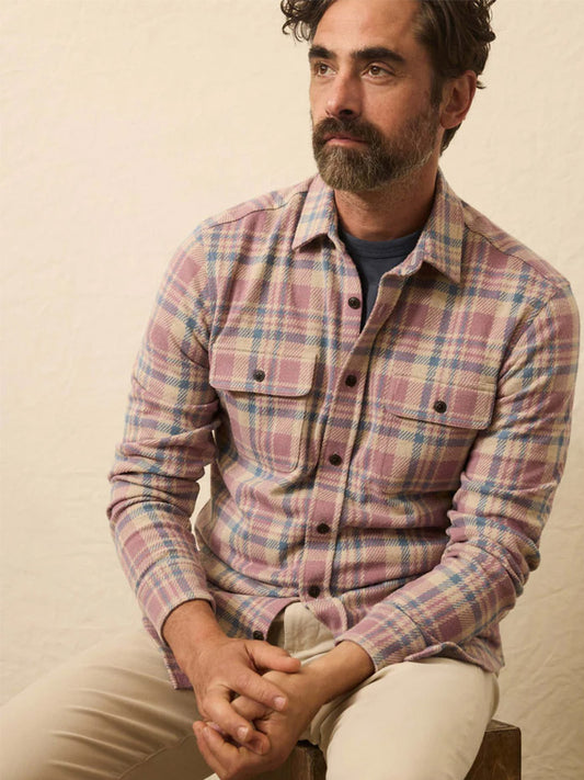 A bearded man sits on a wooden stool, wearing the Faherty Brand Legend Sweater Shirt in Cedar Shore Plaid with light pants, set against a plain, light background.