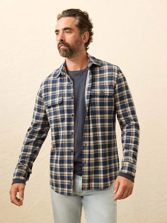 A bearded man wears the Faherty Brand Legend Sweater Shirt in Golden Lake Plaid over a dark t-shirt and light jeans, standing against a neutral background.