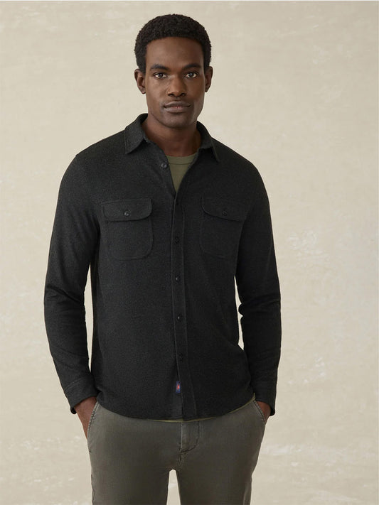 A man stands facing forward in the Faherty Brand Legend Sweater Shirt in Heathered Black Twill, layered over a green T-shirt with gray pants, hands in his pockets against a neutral background.