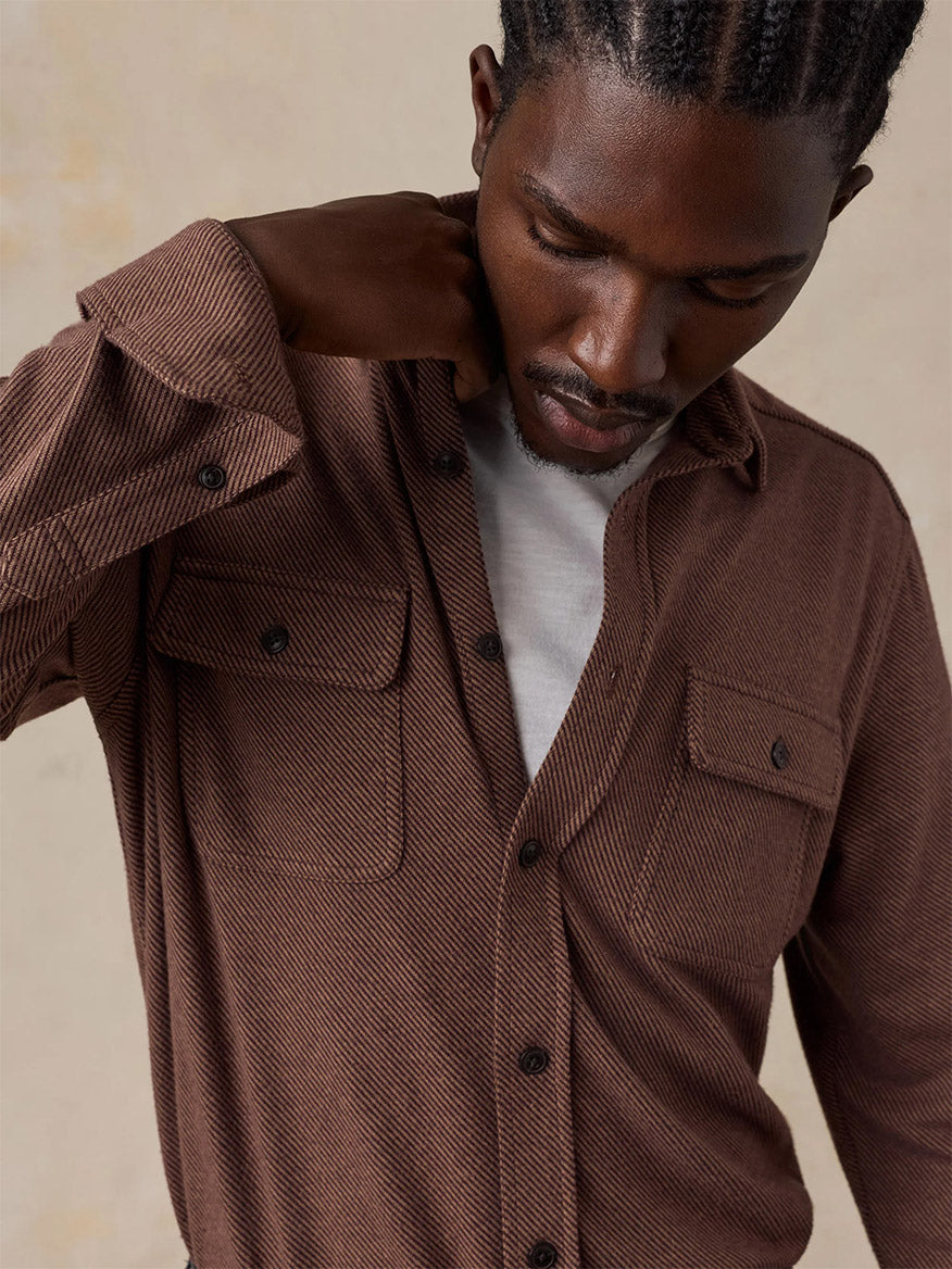 A man wearing a Faherty Brand Legend Sweater Shirt in Redwood Coast Twill over a white t-shirt looks down, pulling at his collar with one hand.